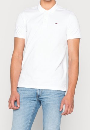 Homme portant un polo blanc à manches courtes avec un petit logo Tommy Hilfiger et un jean bleu clair sur un fond uni.
