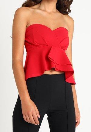 Olkapäät paljastava punainen pepitoppi, jossa on epäsymmetrinen röyhelödetaali, yhdistettynä korkeavyötäröisiin mustiin housuihin, ja henkilö seisoo.