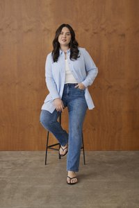 Femme aux longs cheveux foncés assise sur un tabouret contre un mur en bois, portant une chemise bleu clair boutonnée, un haut blanc, un jean bleu et des sandales noires.