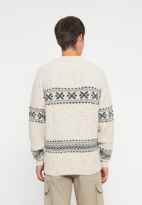 Kreminė megzta palaidinė su pilku geometriniu raštu, ilgomis rankovėmis, apvalia iškirpte ir laisvu siluetu. Textūruotas audinys.