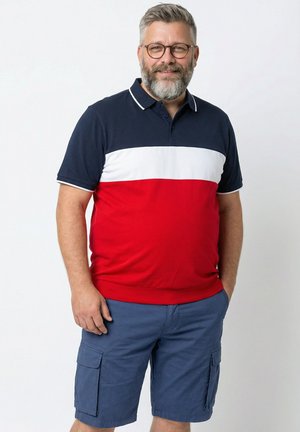 Mężczyzna w średnim wieku z siwymi włosami i brodą, noszący okulary, granatowo-biało-czerwoną koszulkę polo oraz niebieskie szorty cargo, stojący z jedną ręką w kieszeni.