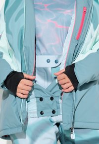 Mátově zelená zimní bunda s růžovými akcenty, s předním zipem a kontrastní vnitřní tkaninou. Převlečená přes vzorovaný top a vyhovující kalhoty.