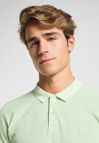 Paita polo hijau muda yang terbuat dari kain tekstur, dengan kerah klasik, placket dua kancing, dan logo bordir kecil di dada.