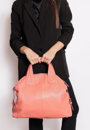 Femme en blazer noir tenant un grand sac à main en cuir corail rose par ses anses, sur fond blanc.