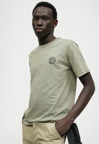 Zielona oliwkowa koszulka bawełniana z okrągłym dekoltem, z małym czarnym logo po lewej stronie na klatce piersiowej. Materiał jest gładki, lekko dopasowany.