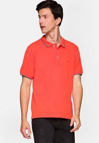Pomarańczowa koszulka polo z kołnierzykiem, z plaketą na trzy guziki oraz kontrastowymi niebieskimi wykończeniami na rękawach. Miękka, gładka bawełniana tkanina.