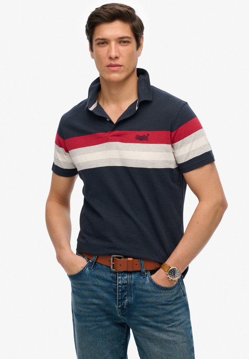 Una camiseta tipo polo azul marino con franjas rojas, blancas y grises en el pecho, que presenta un cuello y mangas cortas. Se lleva con unos pantalones de mezclilla y un cinturón marrón.