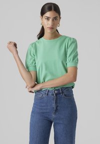 Ljust grönt bomullströja med puffärmar, matchad med högmidjade blå denimjeans. Enkel rundhals och inga mönster.