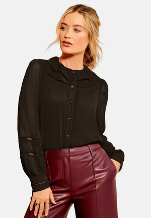 Blonde woman wearing a black blouse with heart-shaped buttons and burgundy leather pants, posing with one hand in pocket.