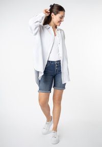 Witte button-up shirt, lichte katoenen textuur, gedragen over een aansluitend wit topje. Donkerdenim shorts met opgerolde zomen, gecombineerd met witte sneakers.