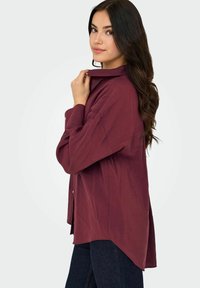 Giovane donna con lunghi capelli scuri, che indossa una camicia button-up bordeaux larga e jeans scuri, in posa di lato con la mano vicino al colletto.