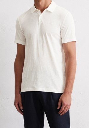 Man die een effen wit poloshirt met korte mouwen en een zwarte broek draagt, staat tegen een witte gestructureerde muur.