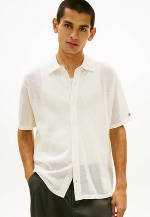 Junger Mann, der ein kurzärmeliges weißes, durchscheinendes Hemd mit schwarzen Knöpfen und schwarze Hose trägt, mit einer Hand in der Tasche stehend, vor einem schlichten Hintergrund.