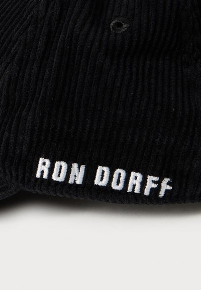 Black ribbed fabric with white embroidered text "RON DORFF" near a metal eyelet on a textured surface.
