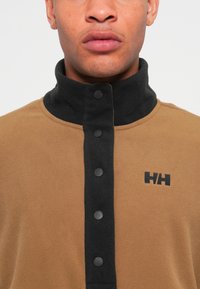 Pull en polaire marron avec un col noir, présentant une patte de boutonnage et un logo noir sur la poitrine. Texture lisse et coupe ajustée.