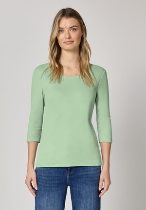 Miętowa bluzka z rękawem do trzech czwartych, wykonana z miękkiego materiału, z zaokrąglonym dekoltem, zestawiona z niebieskimi dżinsami. Prosty fason, dopasowany krój.