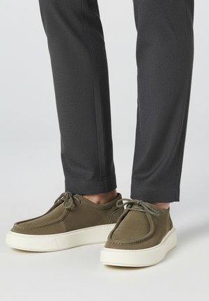 Zapatillas verdes oliva hechas de tela suave, con puntera redonda, cordones atados y suela gruesa de goma blanca; mostradas junto a pantalones grises.