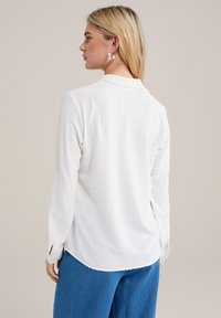 Camicia bianca a maniche lunghe con motivo testurizzato, dotata di colletto e spacchi laterali, abbinata a pantaloni blu a gamba larga.