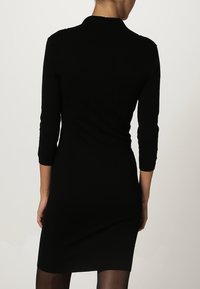 Abito nero dalla forma aderente e maniche a tre quarti. Tessuto a coste al colletto e una silhouette snella che si estende fino a metà coscia.