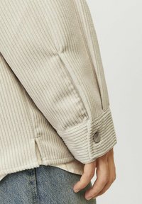 Light beige corduroy jacket sleeve with ribbed texture and buttoned cuff detail. Visible stitching and slight color variation near fabric.