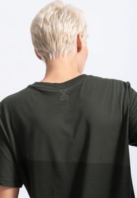 Donkergroene katoenen t-shirt met korte mouwen, met een subtiele schaarlogo op de achterkant, die een zachte textuur en een losse pasvorm laat zien.