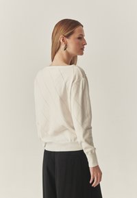 Krém színű pulóver gyémántmintával és bordás mandzsettával. Fekete nadrággal viselve, laza szabású és széles nyakkivágású.