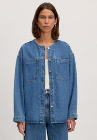 Blaue Jeansjacke mit rundem Ausschnitt, Knopfvorderseite, zwei Brusttaschen und entspannter Passform. Der Stoff hat eine klassisch ausgewaschene Textur.