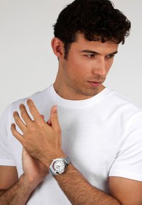 Orologio in metallo argentato con quadrante bianco e accentuazioni rosse. Modello che indossa una maglietta bianca semplice e mostra l'orologio al suo polso.