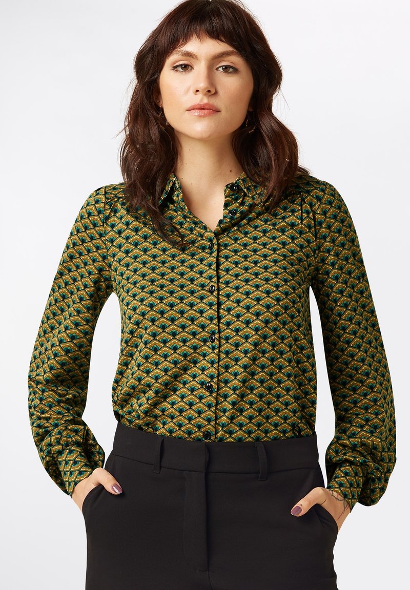 Bluse in grün-gelbem Muster mit Knopfleiste, langen Ärmeln und Kragen, kombiniert mit schwarzen Hosen.