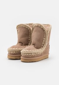 Bottes chelsea en suède beige avec des coutures tressées décoratives le long du bord supérieur, doublure en peluche et semelle en caoutchouc avec une surface texturée.