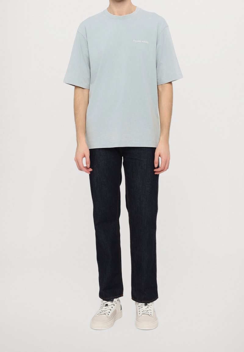 Personne portant un t-shirt ample bleu clair, un jean droit foncé et des baskets blanches, debout devant un fond blanc uni.