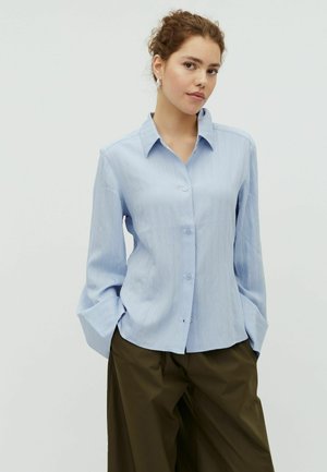 Młoda kobieta o kręconych włosach, ubrana w jasnoniebieską koszulę na guziki i ciemnooliwkowe spodnie typu culotte, stojąca z rękami w kieszeniach.