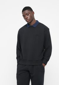 Felpa nera con colletto blu marino, caratterizzata da una vestibilità rilassata, polsini a coste e un piccolo logo sul petto. Indossata con pantaloni neri coordinati.