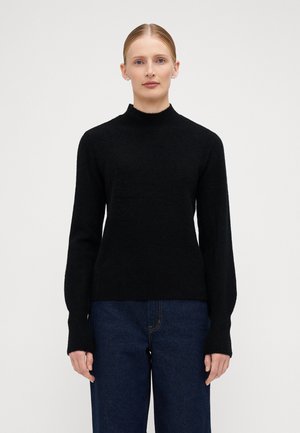Black, fitted sweater with a high neck and long sleeves, made from a soft, textured knit fabric. Paired with dark blue jeans.