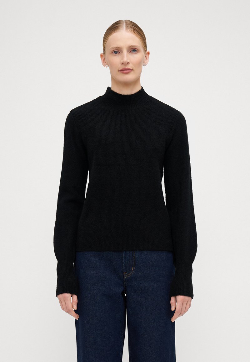 Maglione nero aderente con collo alto e maniche lunghe, realizzato in un morbido tessuto a maglia texturizzato. Abbinato a jeans blu scuro.