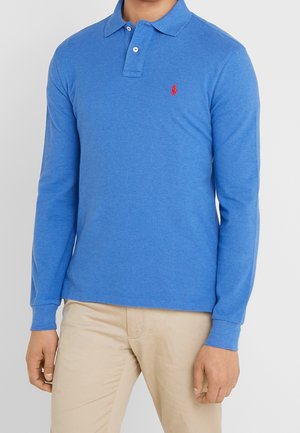 Homme portant un polo bleu à manches longues avec un logo rouge sur la poitrine, associé à un pantalon beige, debout devant un fond uni.