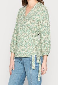 Bloemrijke wikkeltop in groen en crèmekleur, met lange pofmouwen en een gestrikte taille. Gemaakt van een lichte stof met een gestructureerd patroon.