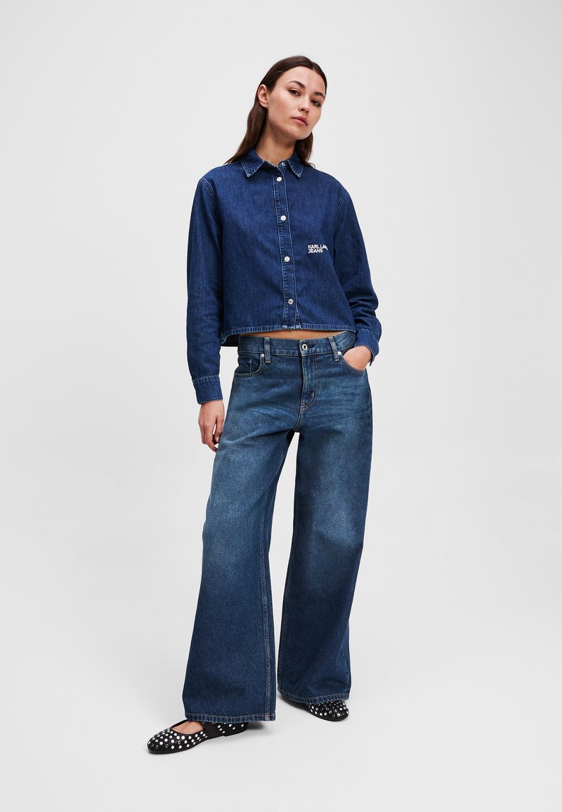 Denim zkrácená bunda s knoflíkovým zapínáním a vyšívaným logem, spárovaná s volnými modrými džíny a balerínkami s puntíky.