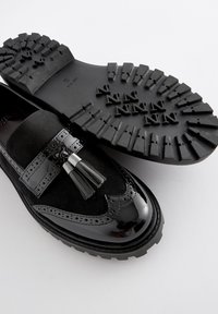 Schwarze Loafers mit einem Obermaterial aus Wildleder und Akzenten aus Lackleder, ausgestattet mit einer Quastenverzierung, einem perforierten Design und einer strukturierten schwarzen Gummisohle.