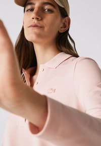 Šviesiai rožinė polo marškinėliai su apykakle, su dviem mygtukais ir išsiuvinėta logo ant kairiojo krūtinės. Glotni tekstūra, ilgomis rankovėmis.