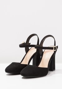 Černé semišové lodičky na vysokém podpatku se čtveratou špičkou, řemínkem kolem kotníku a zlatými kovovými doplňky. Silné blokové podpatky poskytují stabilní základ.