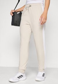 Béžové tepláky s bielymi pruhmi na bočných stranách, elastickým pásom a manžetami. V páre s bielymi športovými topánkami s čiernymi akcentami.