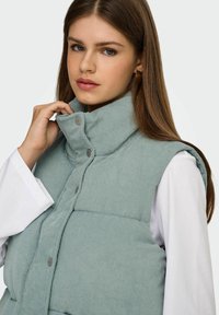 Giovane donna che sistema il colletto di un gilet imbottito verde chiaro sopra una camicia bianca a maniche lunghe, su uno sfondo neutro.