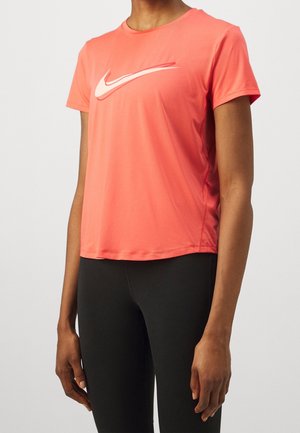 Person trägt ein korallenfarbenes Nike-T-Shirt mit kurzen Ärmeln und weißem Logo, kombiniert mit schwarzen, eng anliegenden Hosen, stehend vor einem hellen Hintergrund.