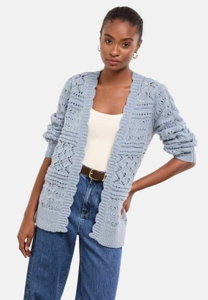 Light blue knitted cardigan with a lace pattern, open front, and ribbed edges. Long sleeves and relaxed fit. Worn over a cream top.