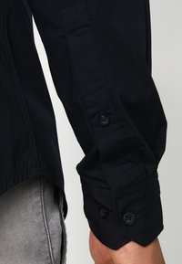 Ciemnoniebieska koszula z długim rękawem z guzikami na mankietach, o gładkiej teksturze materiału i dopasowanym kroju.