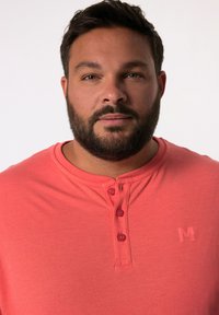 Korálová krátká rukávová henley košile se třemi knoflíky na límci. Obsahuje jemné logo na hrudi. Měkká látka a uvolněný střih.