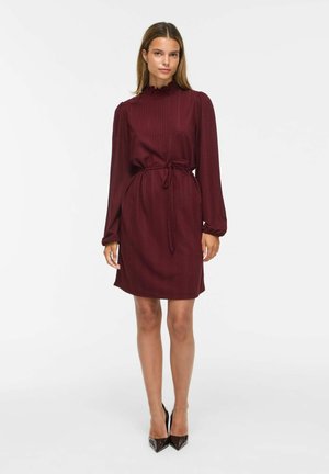 Femme portant une robe bordeaux à manches longues, longueur genou avec taille nouée et des talons noirs à bout pointu, debout devant un fond uni.