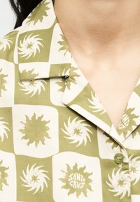 Πουκάμισο με γιακά σε πράσινο και λευκό καρό μοτίβο που παρουσιάζει στυλιζαρισμένα ηλιακά σχήματα, ένα από τα οποία περιέχει το κείμενο «Santa Cruz».