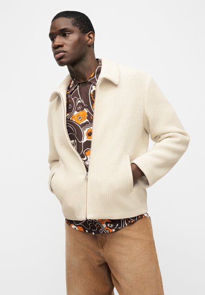 Άνδρας που φοράει κρεμ πλεκτό μπουφάν με φερμουάρ πάνω από καφέ πουκάμισο με λουλούδια και ανοιχτό καφέ παντελόνι, τα χέρια στις τσέπες του μπουφάν, κοιτάζοντας προς τα αριστερά.